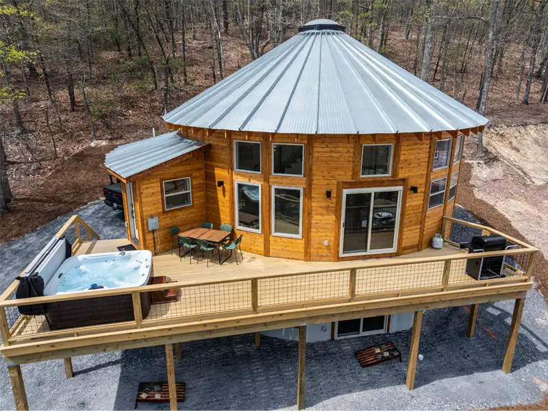 A round house with a hot tub on the roof, nestled in a natural setting with greenery and a bright sky.